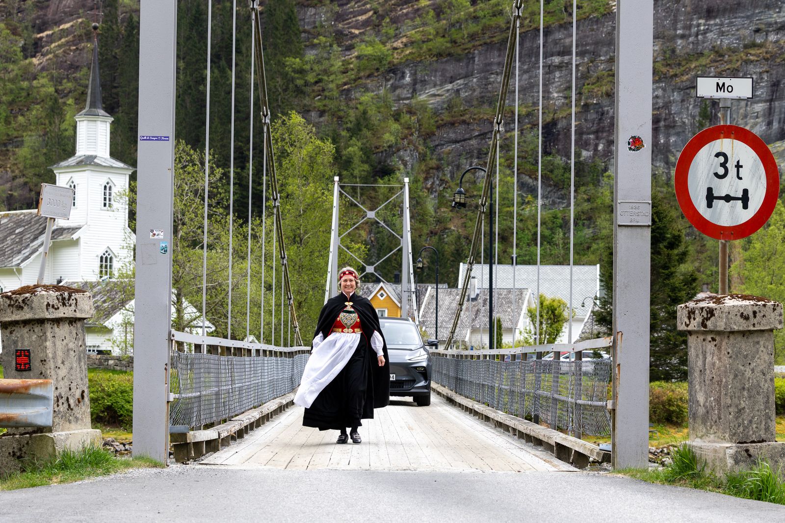 En kvinne i svart og rød bunad med hvitt skjørt og rød hodepryd går over en hengebro mot kamera. I bakgrunnen ses en hvit kirke, bolighus og en bil på broen.