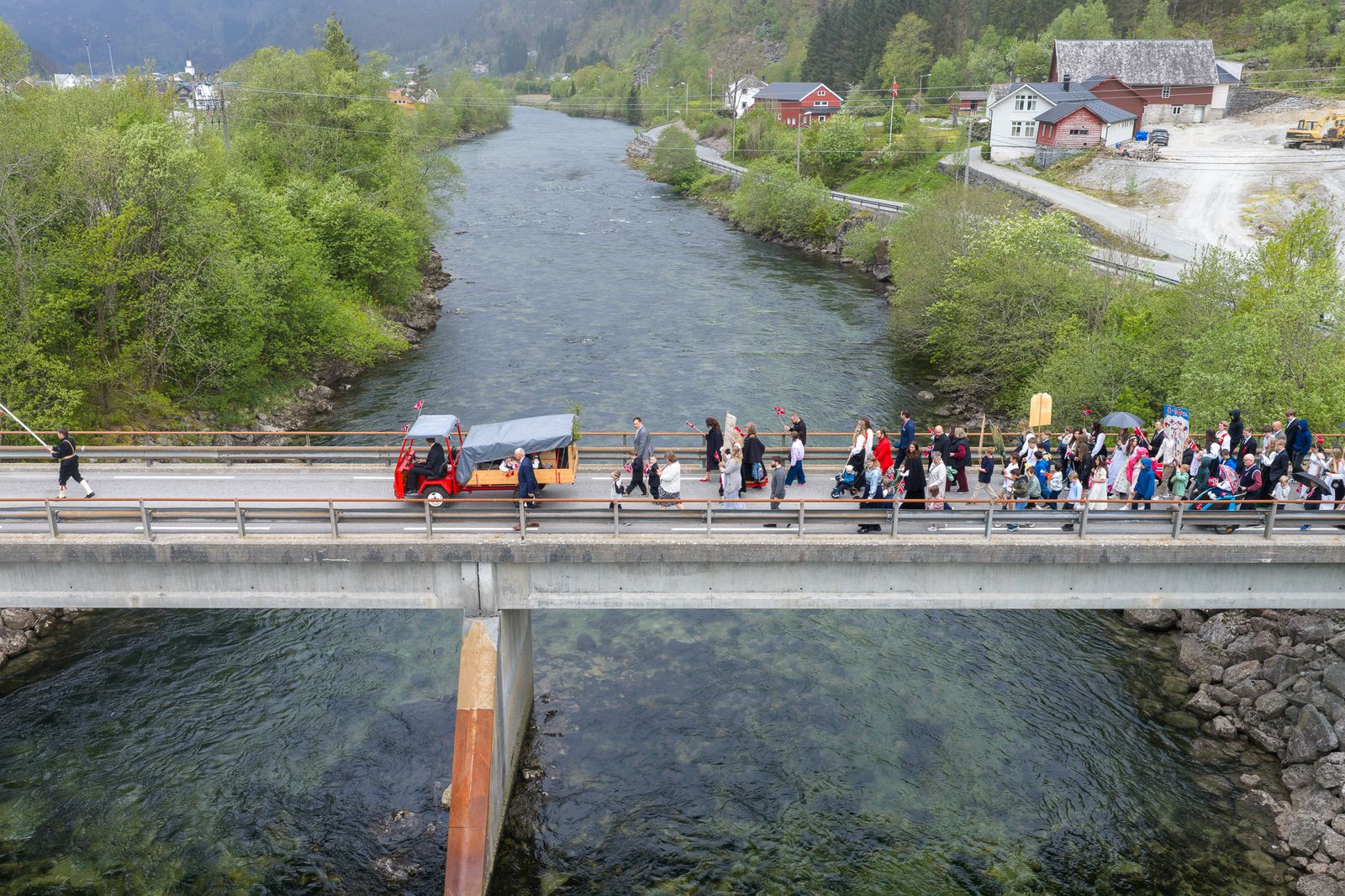 Dronebilder tatt ovenfra viser en folkemengde som går i tog over en betongbro over en elv. En rød trehjulet elektrisk lastesykkel leder an i prosesjonene. Deltakerne holder norske flagg og er kledd i mørke antrekk og hvite kjoler. Grønne åssider og norske bygninger ses i bakgrunnen.