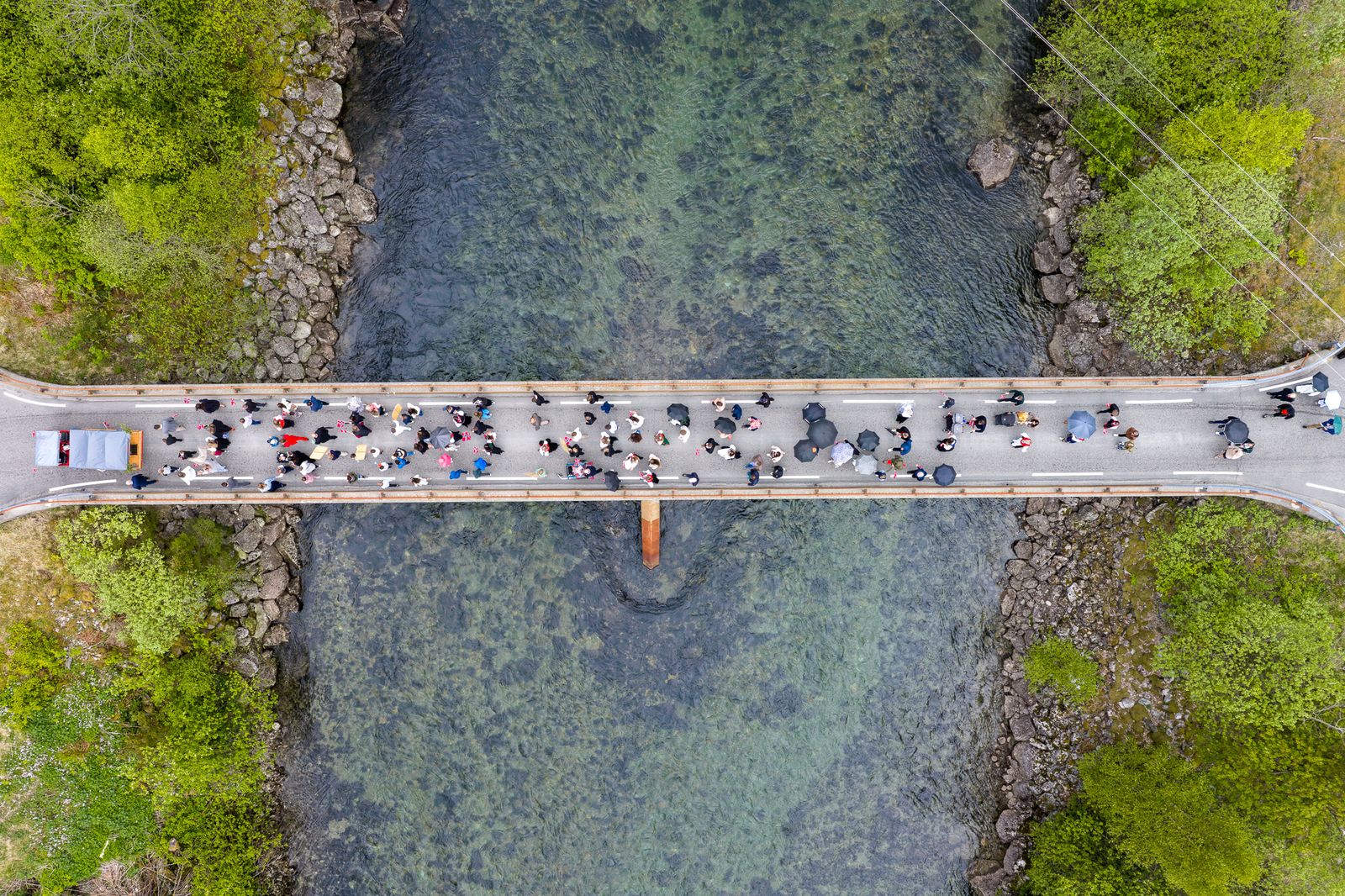 Dronefoto rett ovenfra av en bro over en grønn elv. En folkemengde på anslagsvis 60–80 personer går i rekke langs broen. Flere holder paraply. På venstre side av broen står et mindre kjøretøy med grå presenning. Grønn vegetasjon og rullestein langs elvekantene på begge sider.