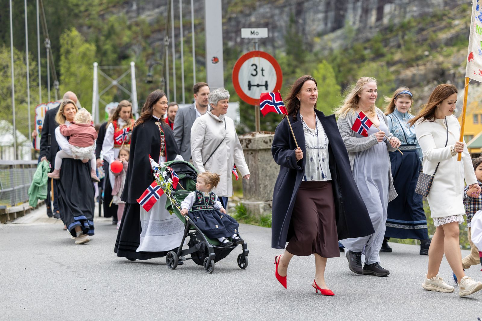 En gruppe mennesker, flere i bunad og tradisjonelle drakter, går i tog over en bro. En kvinne i mørk frakk og røde hæler holder et norsk flagg, en annen triller en barnevogn med et lite barn i bunad. I bakgrunnen ses en fjellside med grønn vegetasjon.