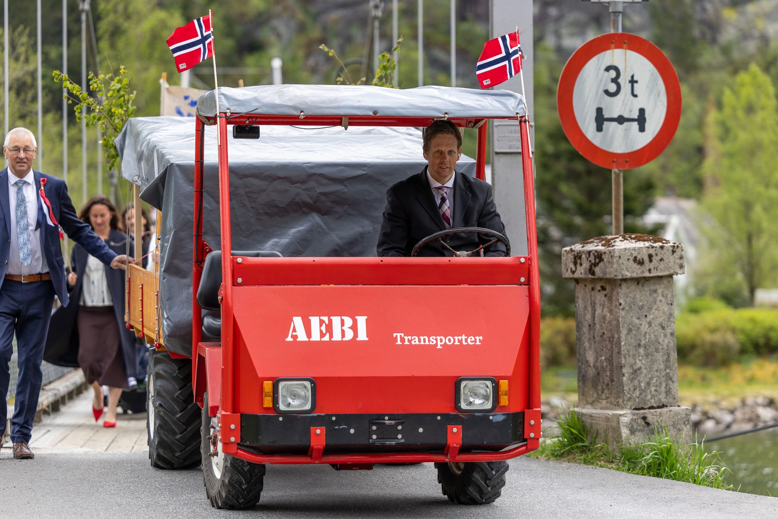 En mann i mørk dress kjører en rød AEBI Transporter med to norske flagg på taket over en bro. Bak ham følger flere voksne personer til fots. Ved siden av veien står et skilt med maksimal aksellast 3 tonn.